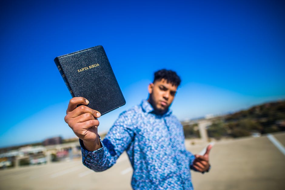 A young man with a floral shirt holding a 'Santa Biblia' against a clear blue sky.